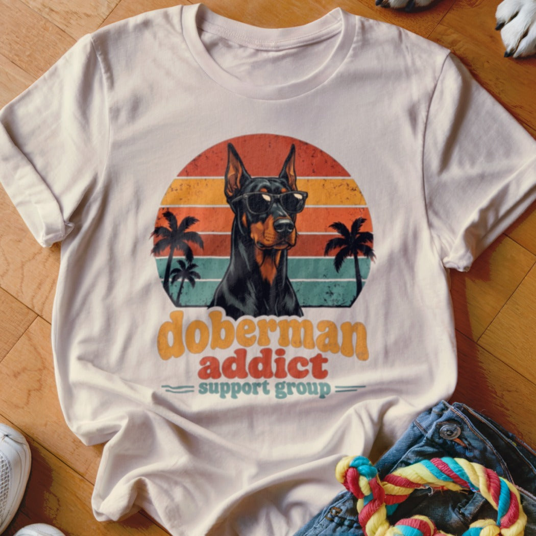 Beige t-shirt featuring a retro sunset graphic of a Doberman wearing sunglasses and palm trees.
