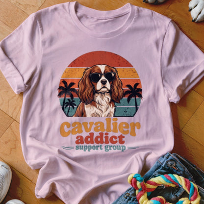 A light pink t-shirt with a vintage-style graphic of a Cavalier King Charles Spaniel wearing sunglasses in front of a sunset.