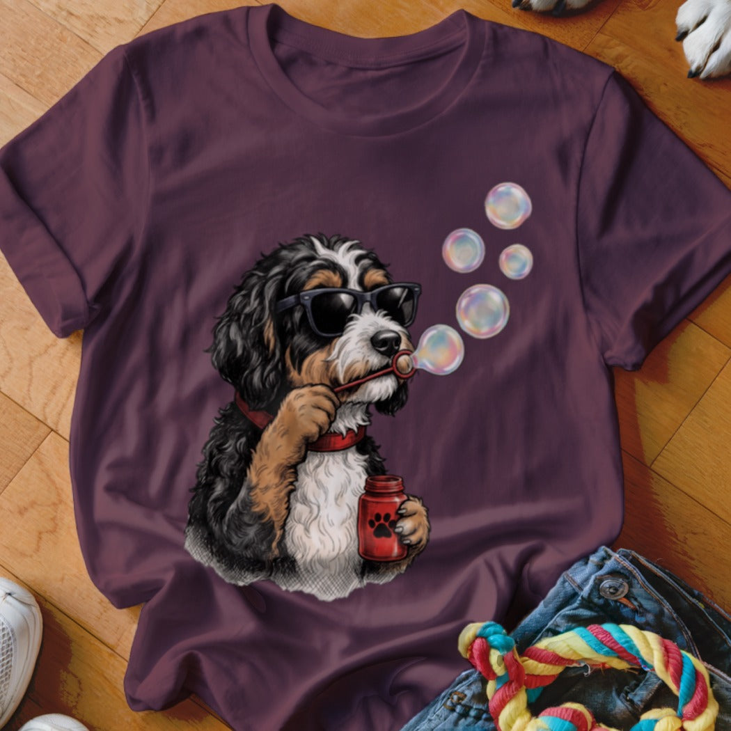 A dark purple t-shirt features a graphic of a Bernedoodle wearing sunglasses and blowing bubbles.