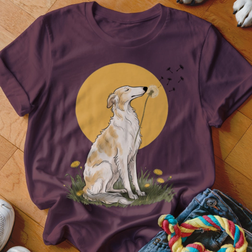 Maroon t-shirt featuring an illustration of a dog sitting in a field blowing dandelions against a yellow sun.
