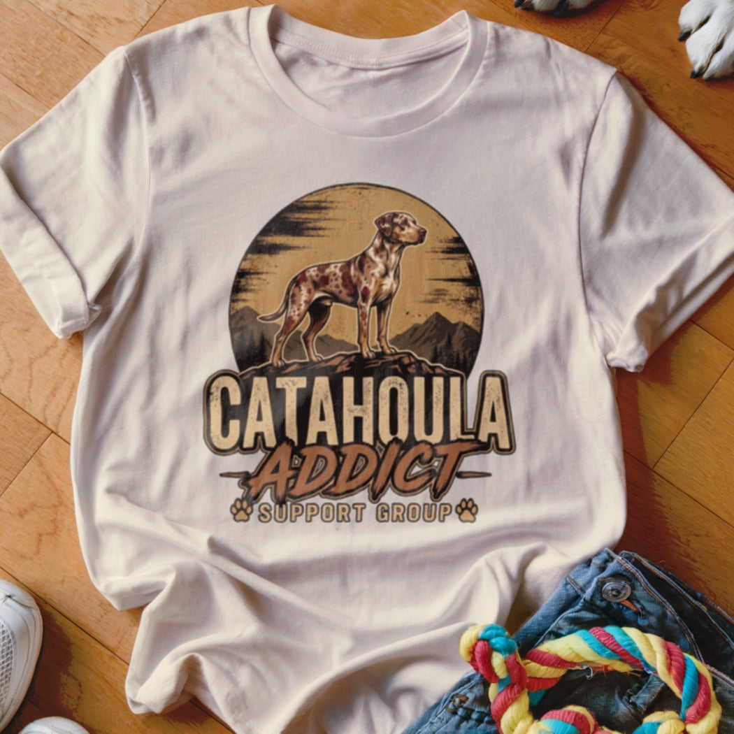 A light-colored t-shirt featuring a retro graphic of a Catahoula leopard dog standing on mountains.
