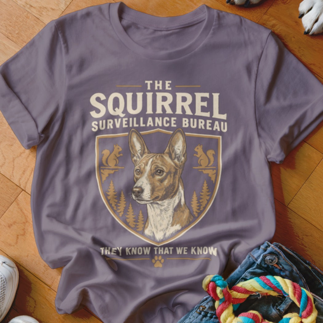 Purple t-shirt featuring a graphic of a dog, pine trees, squirrels, and text reading The Squirrel Surveillance Bureau.