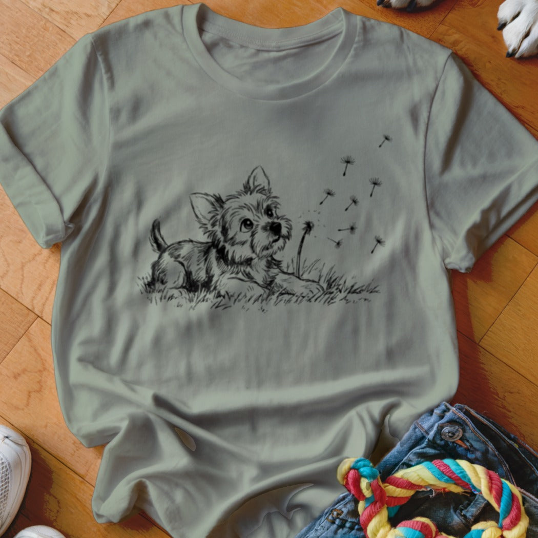 Sage green t-shirt featuring a black line drawing of a puppy watching dandelion seeds blow away.