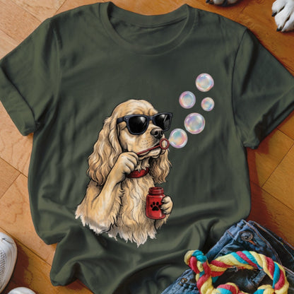An olive green t-shirt featuring a graphic of a cocker spaniel wearing sunglasses and blowing bubbles.