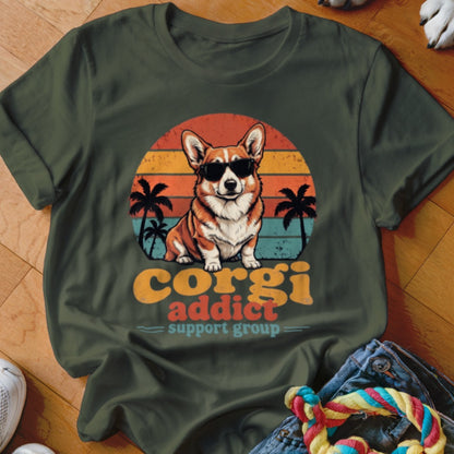 An olive green t-shirt featuring a retro graphic of a corgi wearing sunglasses before a sunset.