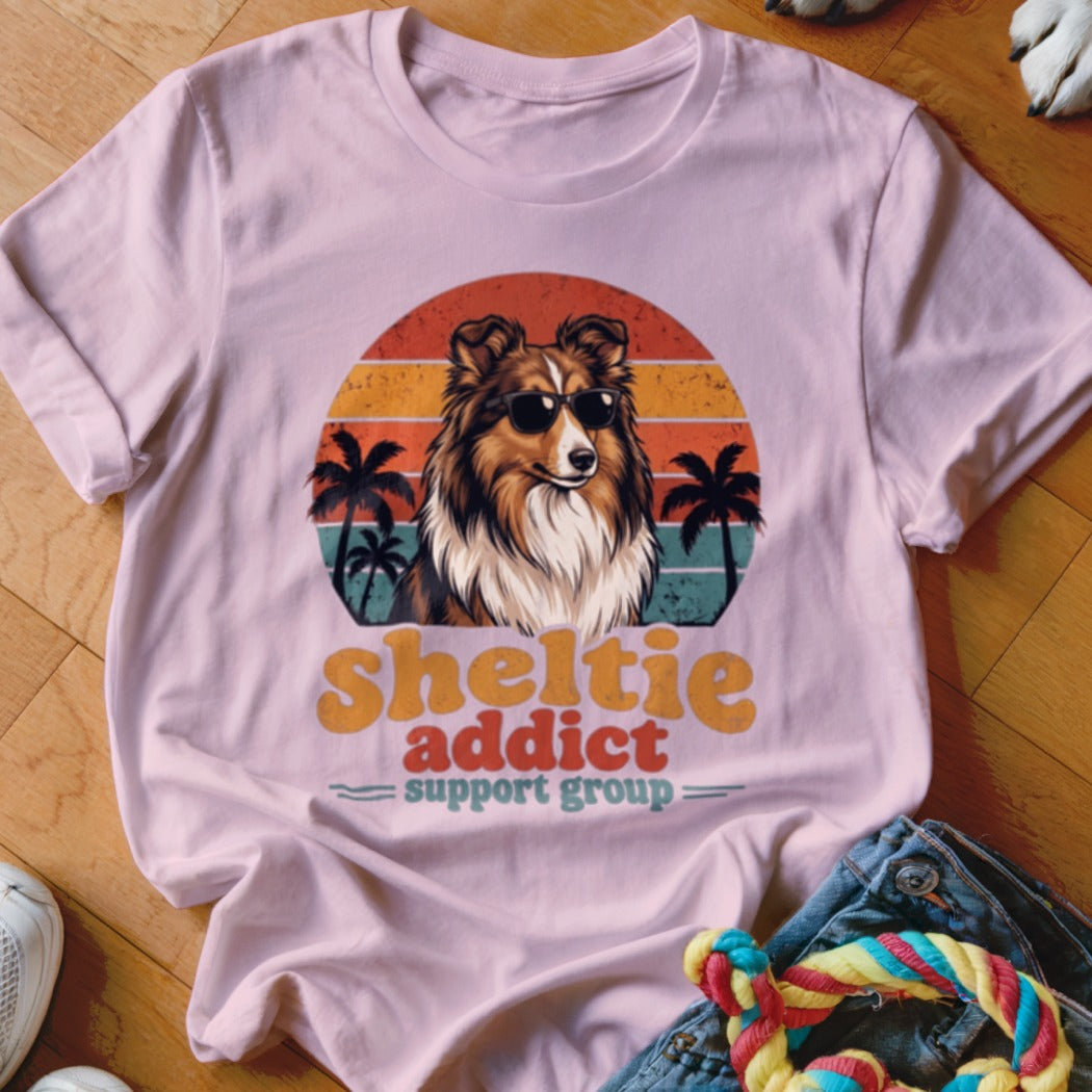 A pink t-shirt featuring a retro Sheltie graphic with sunglasses, palm trees, and text reading Sheltie addict support group.