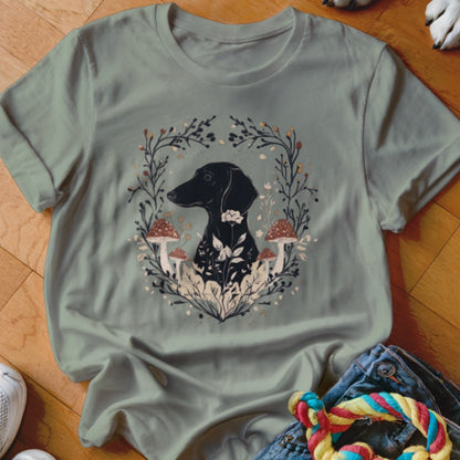 A sage green t-shirt featuring a graphic of a black dachshund surrounded by mushrooms, flowers, and vines.