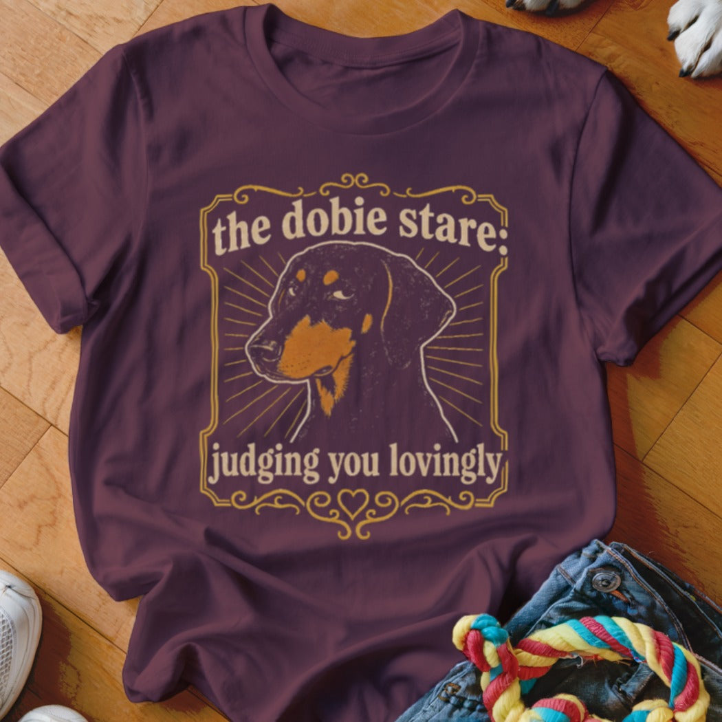 Doberman dog graphic with text "the dobie stare: judging you lovingly" on a purple shirt.