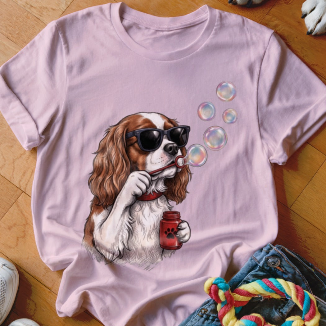 A light pink t-shirt featuring a graphic of a Cavalier King Charles Spaniel wearing sunglasses and blowing bubbles.
