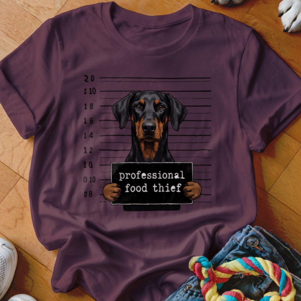 Doberman with "professional food thief" sign against a mugshot background.