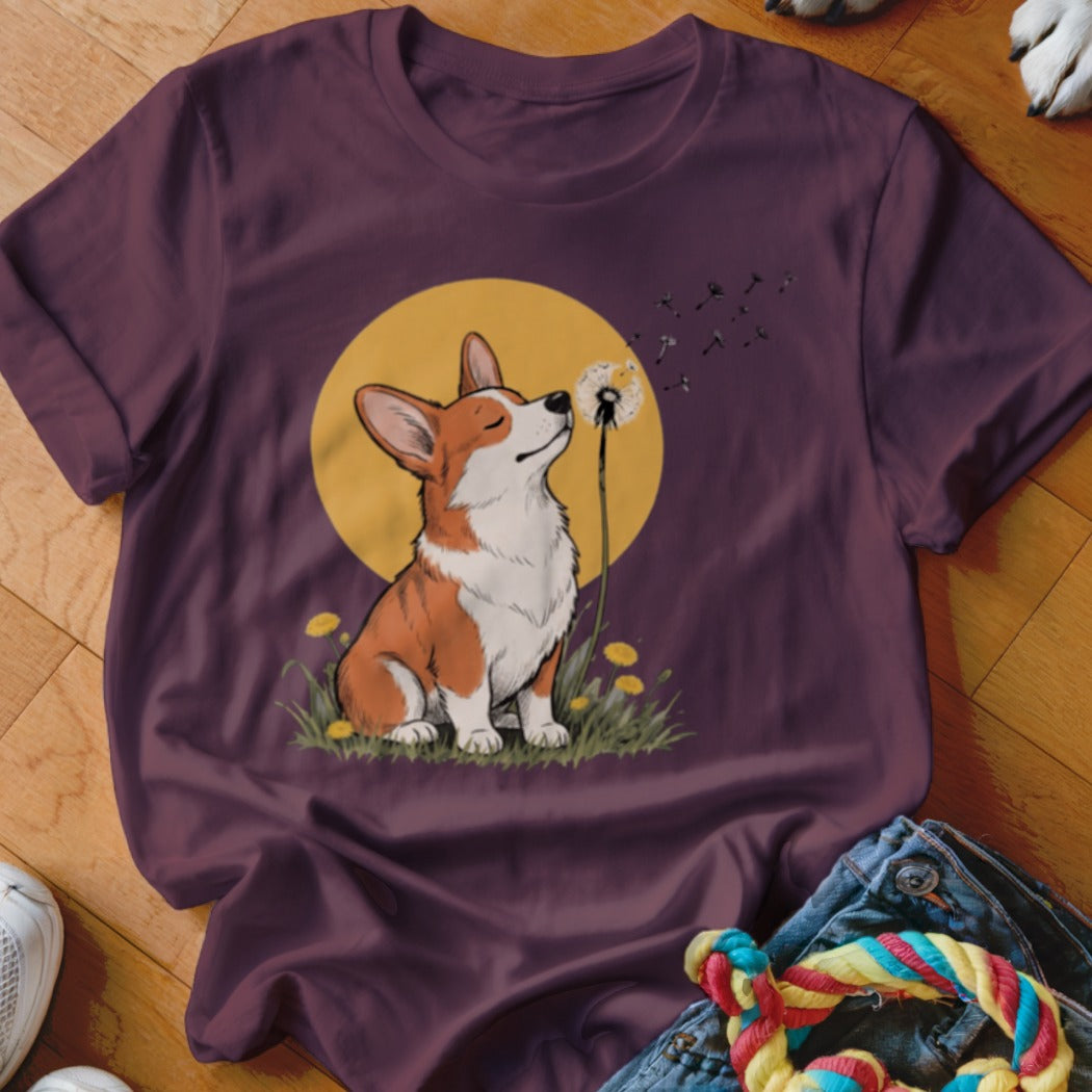 Maroon t-shirt featuring a graphic of a corgi blowing dandelion seeds in front of a yellow sun.
