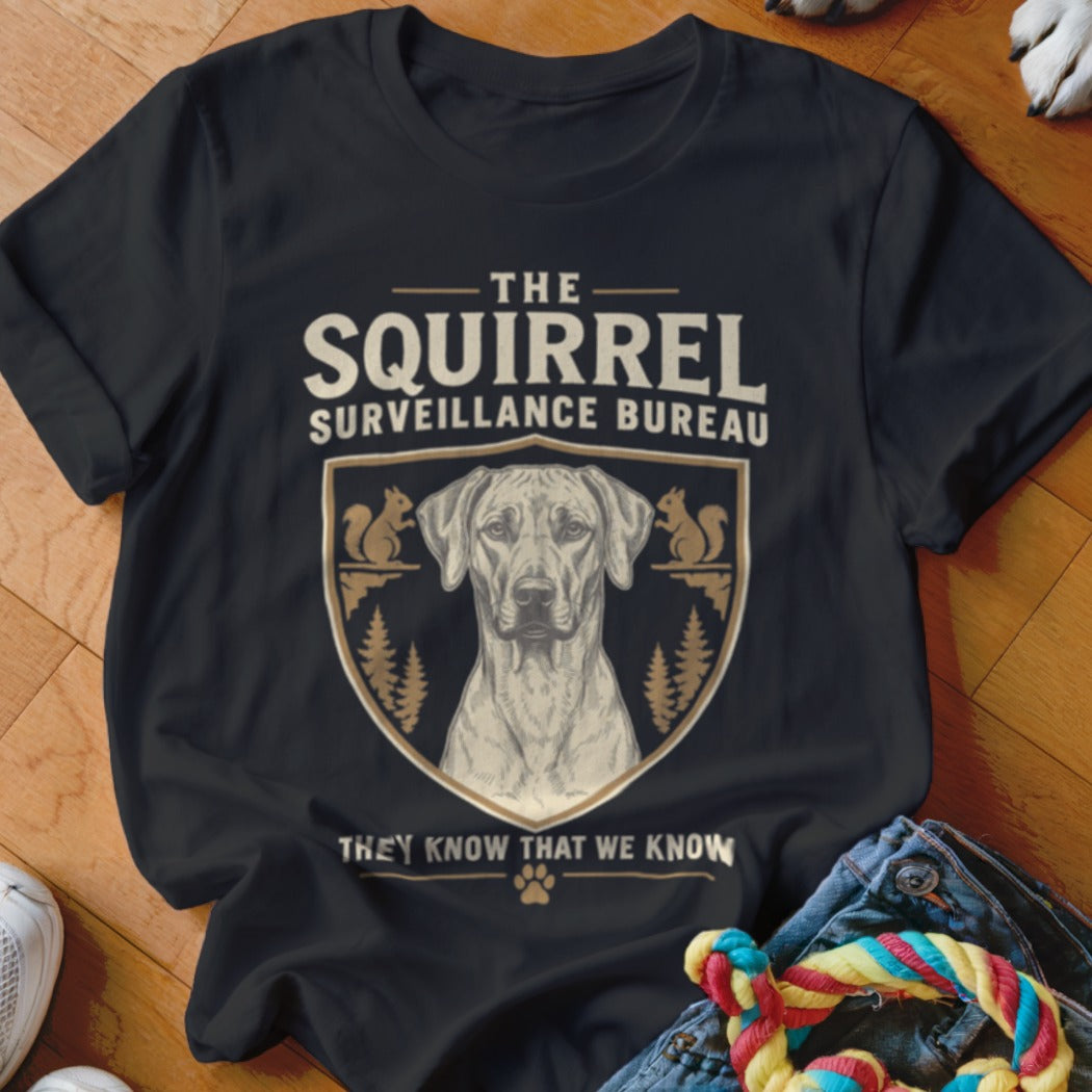 Black t-shirt featuring a crest with a dog, squirrels, pine trees, and the text The Squirrel Surveillance Bureau.