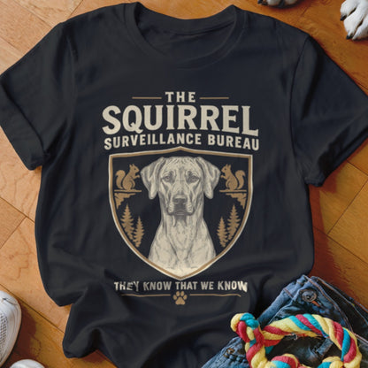 Black t-shirt featuring a crest with a dog, squirrels, pine trees, and the text The Squirrel Surveillance Bureau.