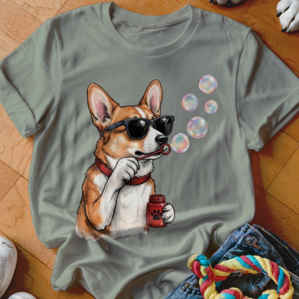 A sage green t-shirt featuring a graphic of a corgi wearing sunglasses and blowing soap bubbles.