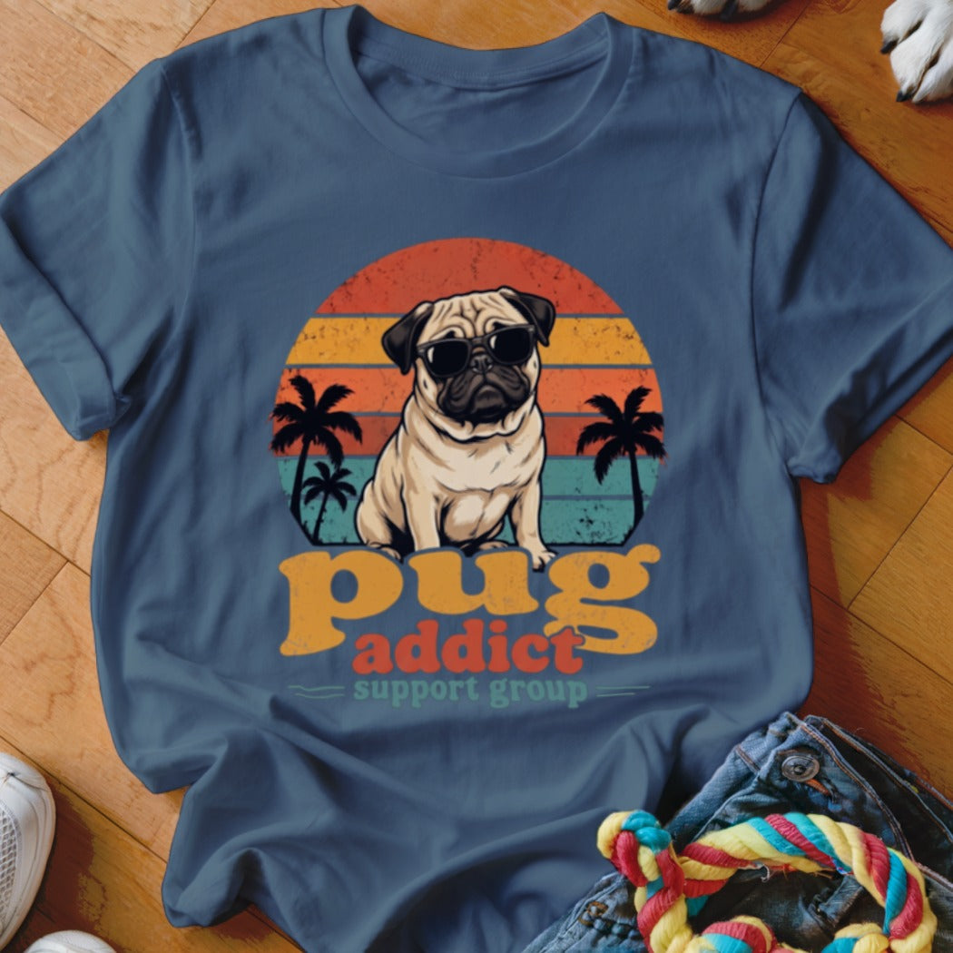 A blue t-shirt featuring a retro graphic of a pug wearing sunglasses in front of a sunset, with palm trees and text reading "pug addict support group".