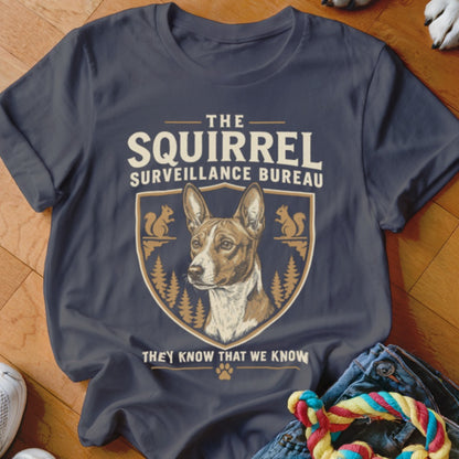 A dark grey t-shirt featuring a graphic of a dog, squirrels, trees, and text reading The Squirrel Surveillance Bureau.