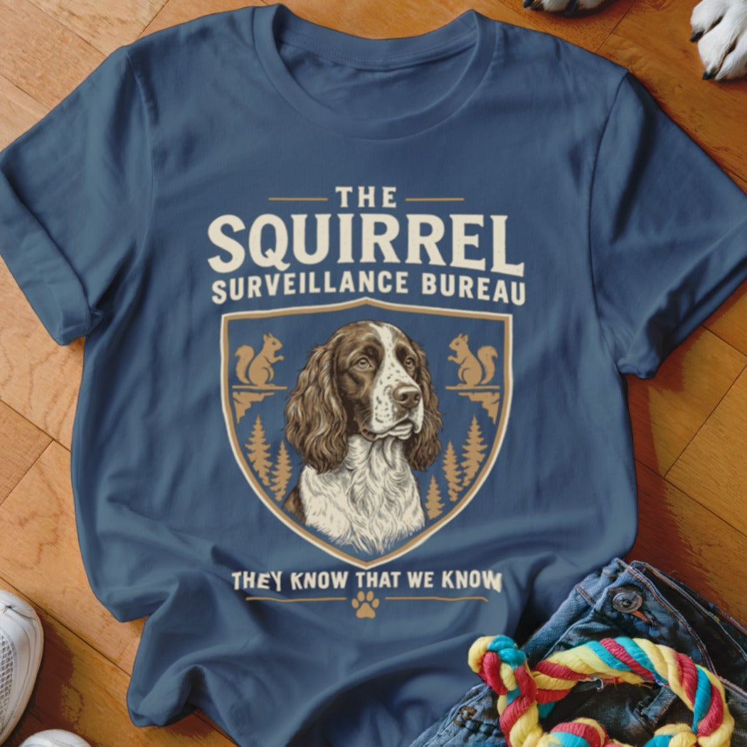 Blue t-shirt featuring a crest with a dog, pine trees, squirrels, and text reading The Squirrel Surveillance Bureau.
