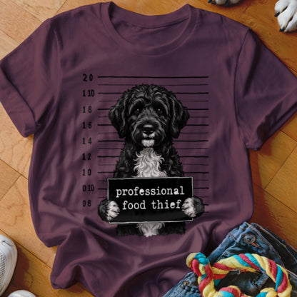 Black dog mugshot with "professional food thief" sign on plum t-shirt.