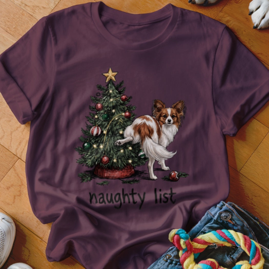 Papillon dog near Christmas tree with text "naughty list" on burgundy shirt.