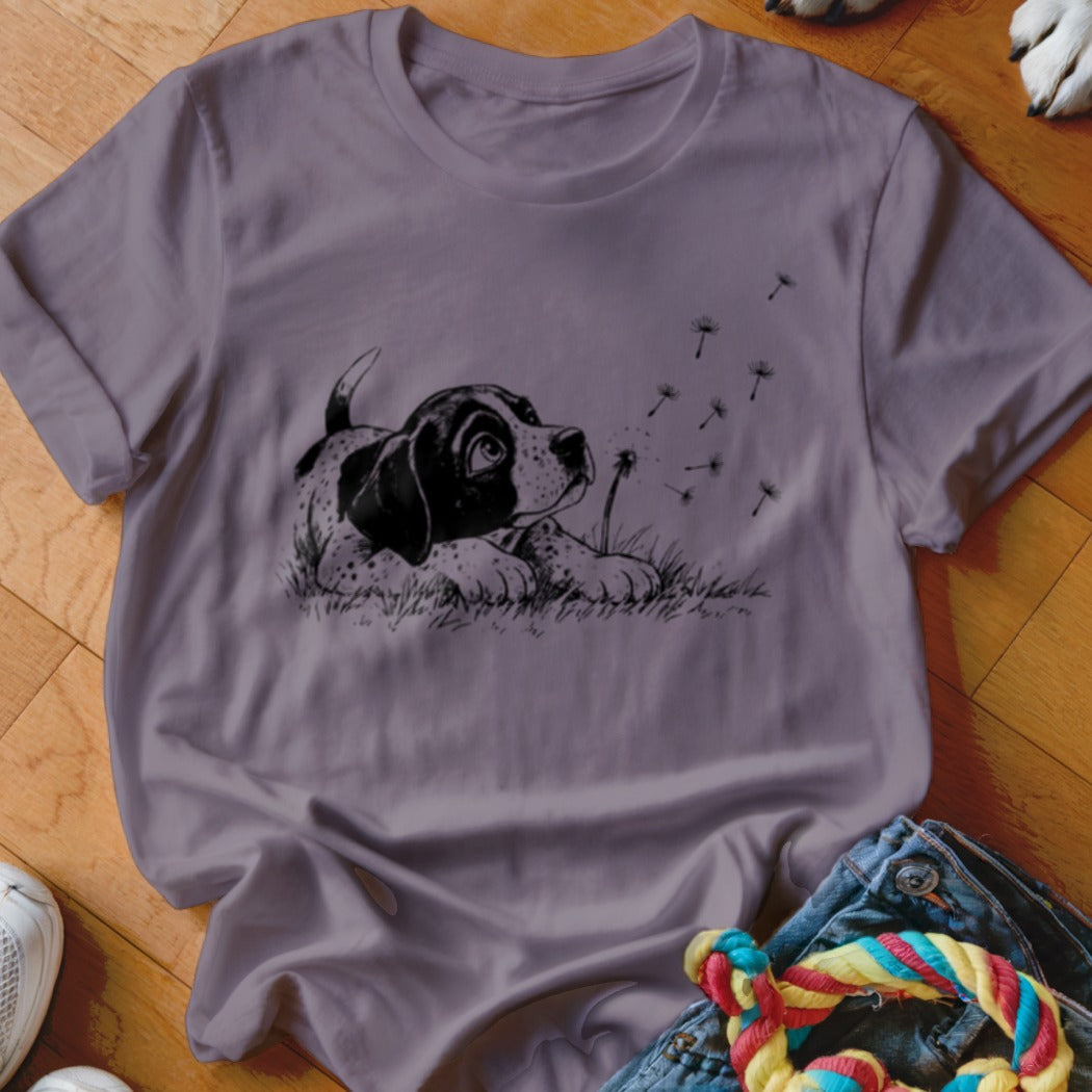 A purple short-sleeved t-shirt featuring a black-and-white graphic of a puppy watching dandelion seeds blow away.
