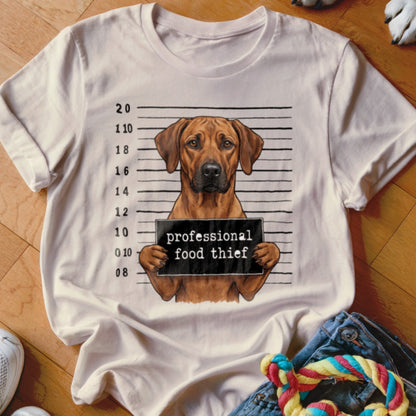 Brown dog mugshot with "professional food thief" sign on a t-shirt.