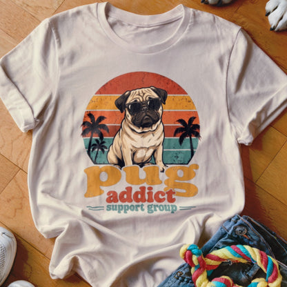 A light-colored t-shirt with a retro graphic of a pug wearing sunglasses in front of a sunset and palm trees, with the text pug addict support group.