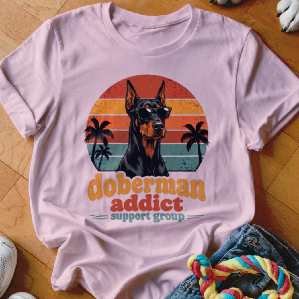Pink t-shirt featuring a retro graphic of a Doberman wearing sunglasses with palm trees, sunset, and text that reads Doberman Addict Support Group.