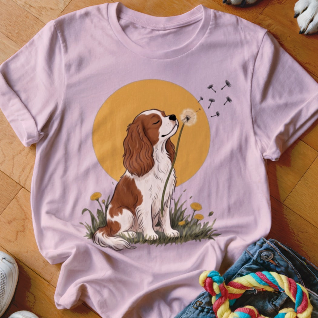 A pink t-shirt featuring an illustration of a Cavalier King Charles Spaniel blowing dandelion seeds.