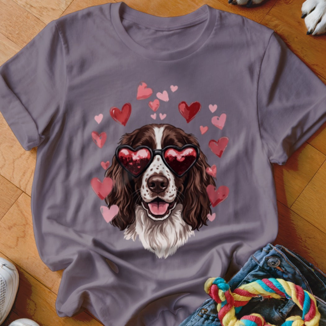 A grey short-sleeved t-shirt featuring an illustration of a brown and white dog wearing heart-shaped sunglasses, surrounded by pink hearts.