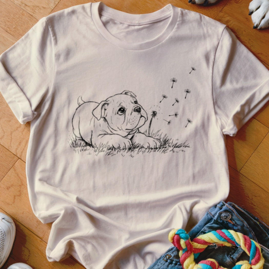 A cream-colored t-shirt featuring a black line art graphic of a bulldog puppy looking at dandelion seeds.