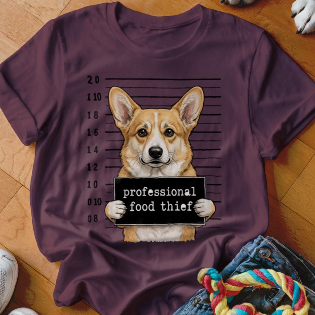 Corgi dog in mugshot holding sign "professional food thief" on maroon shirt.