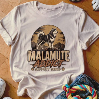 A light beige t-shirt featuring a vintage-style graphic of a Malamute with mountains and text reading Malamute Addict Support Group.