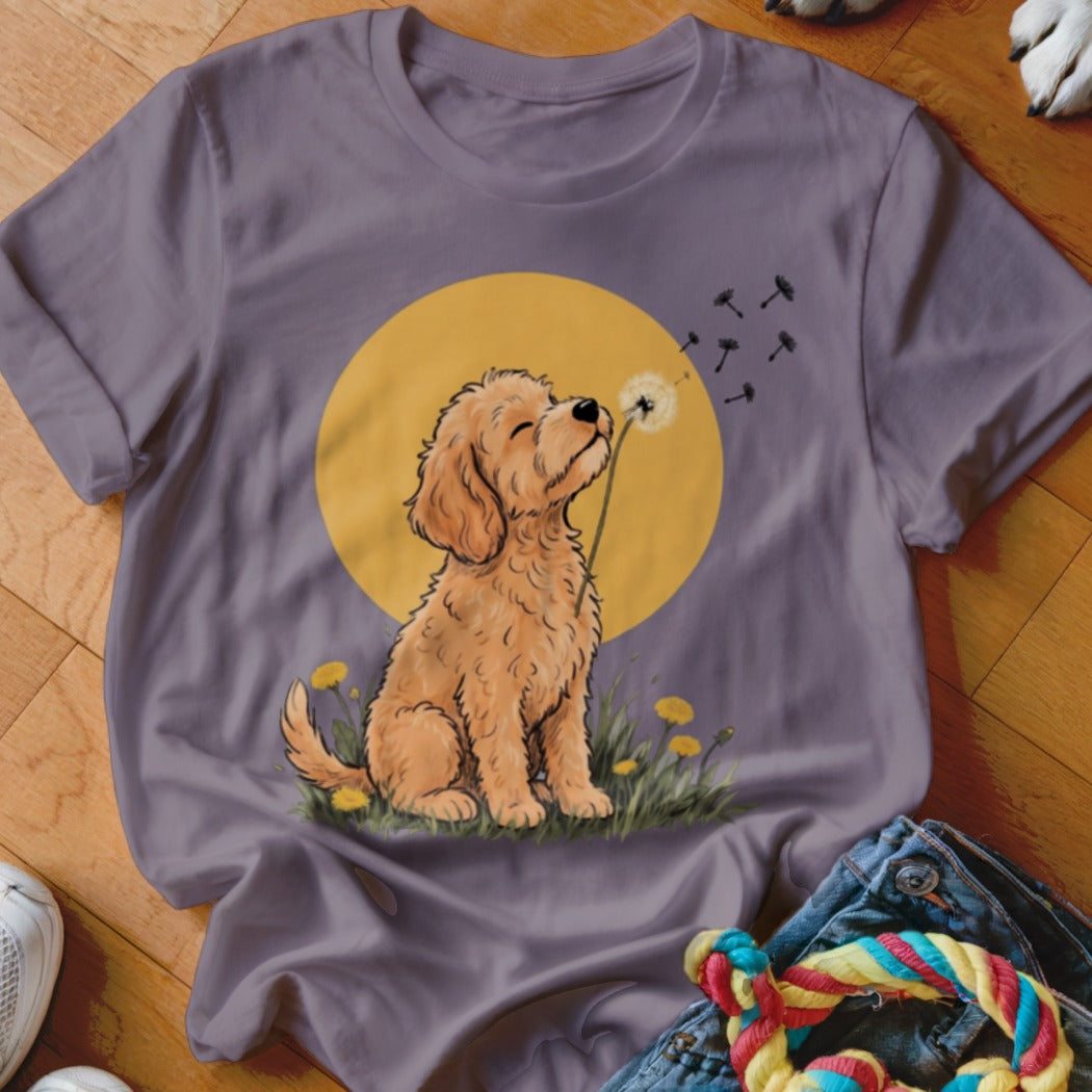 A purple t-shirt featuring an illustration of a fluffy golden dog blowing dandelion seeds.