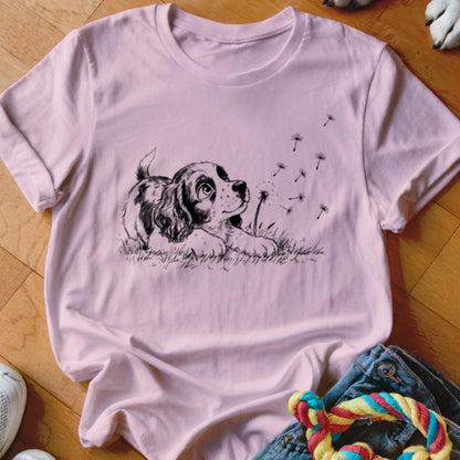 Light pink t-shirt featuring a black graphic of a puppy in the grass watching dandelion seeds blow away.