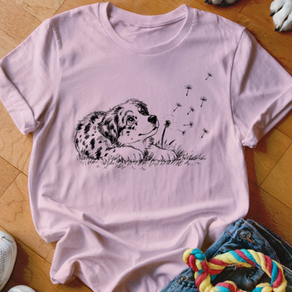 Light pink crewneck t-shirt featuring a black line art graphic of a spotted puppy watching dandelion seeds blow away.