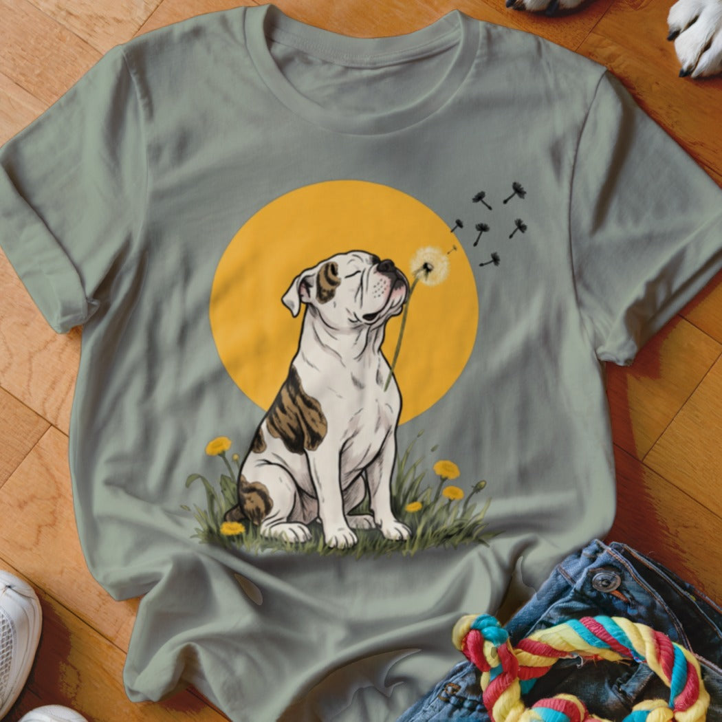 Sage green t-shirt featuring a graphic of a brindle bulldog blowing dandelion seeds against a yellow sun.