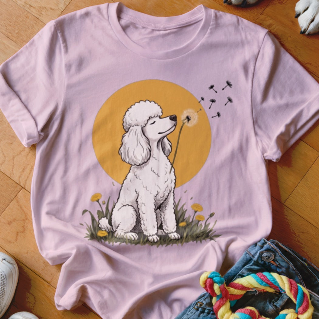 A light pink short-sleeve t-shirt featuring a graphic of a white poodle blowing a dandelion seed head against a yellow sun.