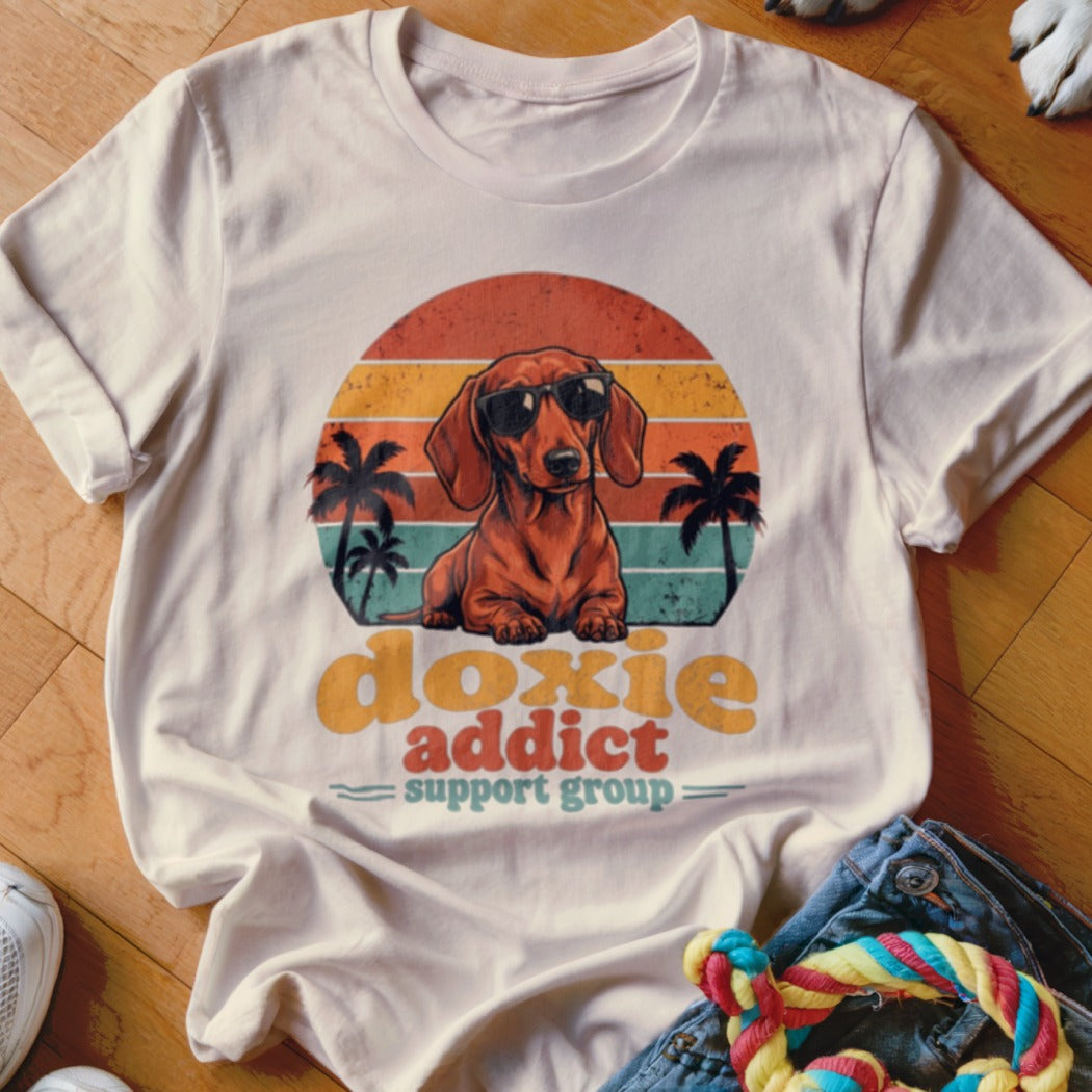 A light beige t-shirt with a vintage-style graphic of a dachshund wearing sunglasses before a sunset.