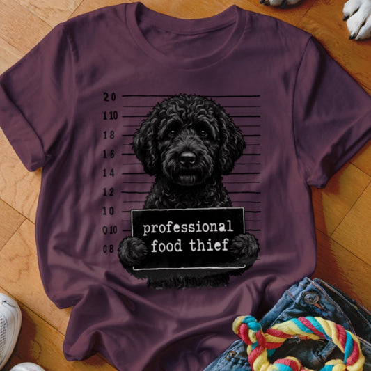 Black poodle in mugshot holding "professional food thief" sign on a maroon t-shirt.