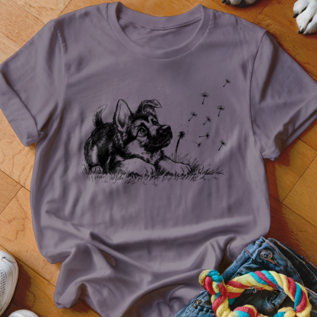 A crewneck t-shirt in muted purple features a black sketch of a puppy watching dandelion seeds blow away.