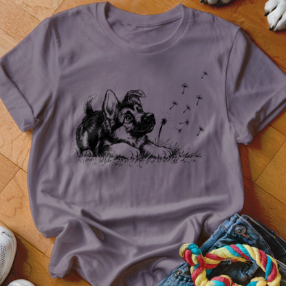 A crewneck t-shirt in muted purple features a black sketch of a puppy watching dandelion seeds blow away.