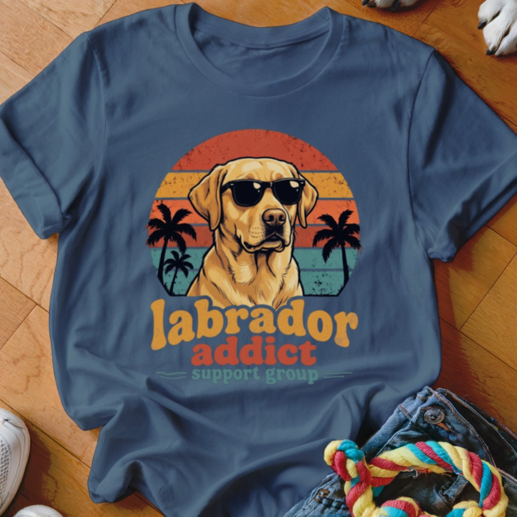 A dark blue t-shirt with a graphic of a yellow Labrador wearing sunglasses before a sunset and palm trees, with the text "labrador addict support group".