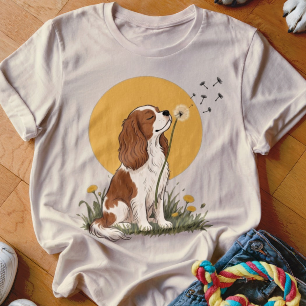 A light beige t-shirt featuring an illustration of a Cavalier King Charles Spaniel blowing dandelion seeds in front of a yellow sun.