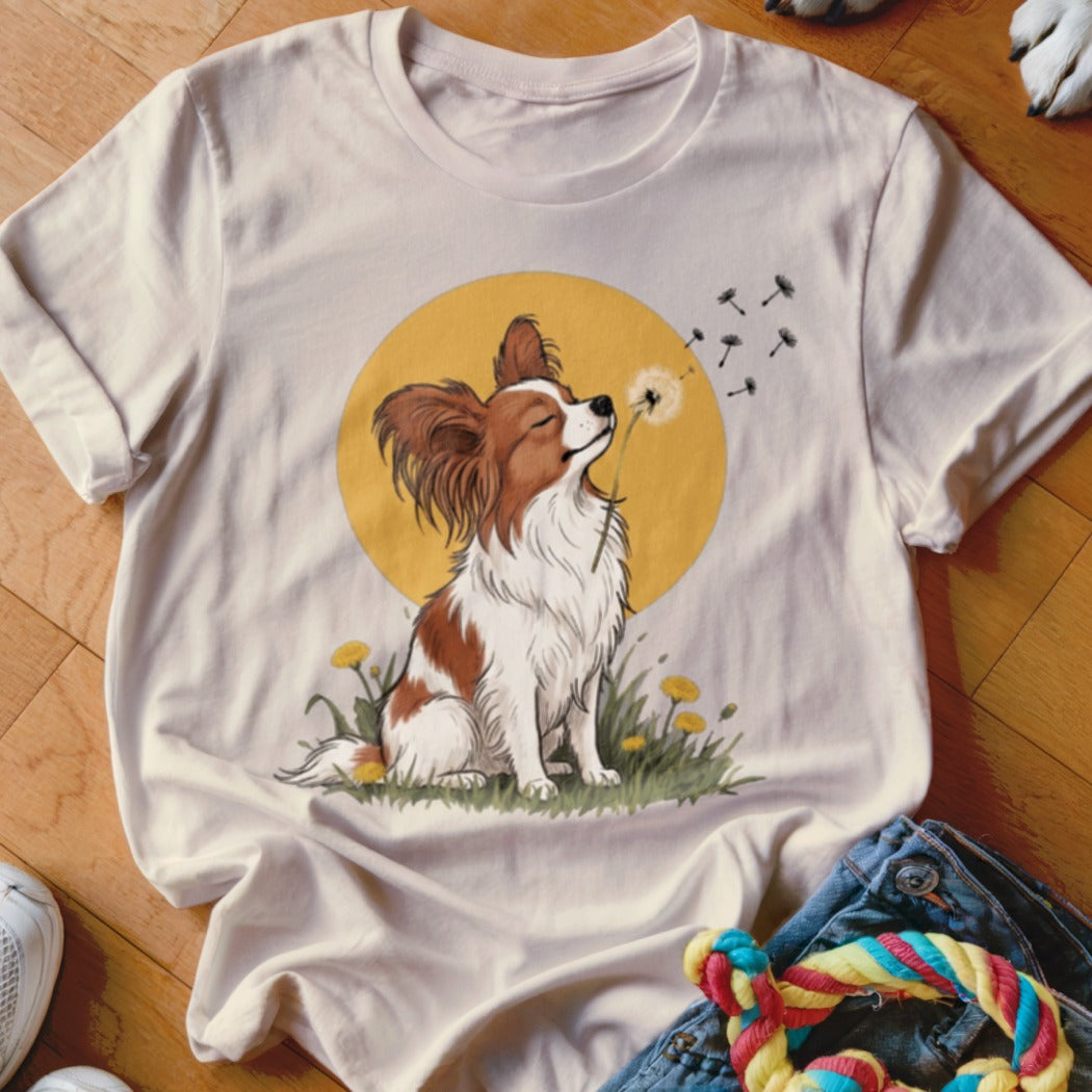 A cream-colored t-shirt with a graphic of a brown and white Papillon dog blowing dandelion seeds against a yellow circle.