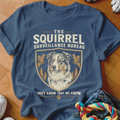 A blue t-shirt printed with a graphic featuring an Australian Shepherd and text reading The Squirrel Surveillance Bureau.