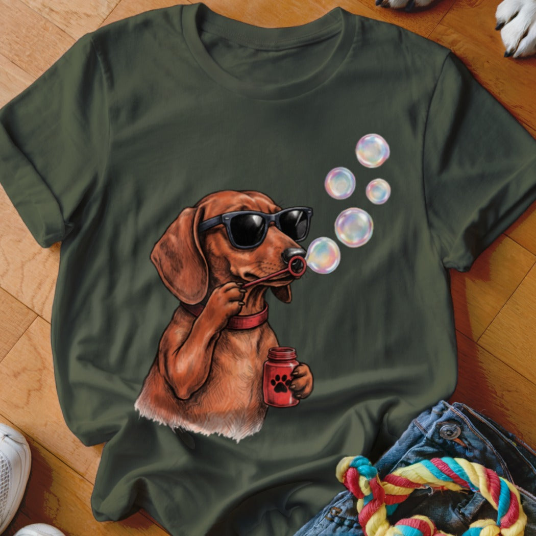 An olive green t-shirt featuring a graphic of a dachshund wearing sunglasses and blowing bubbles.