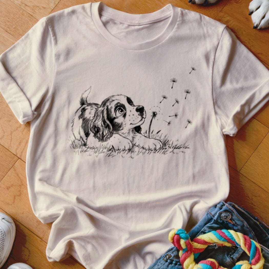 Light pink crewneck t-shirt featuring a black line art graphic of a puppy watching floating dandelion seeds.