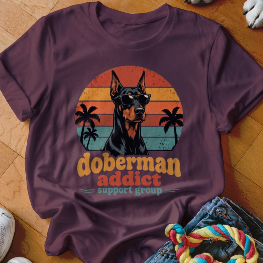A maroon t-shirt featuring a retro graphic of a Doberman wearing sunglasses in front of a sunset.