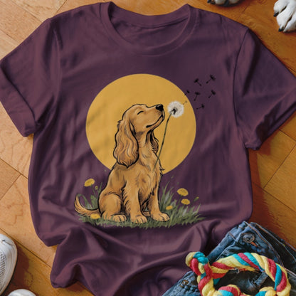 A plum-colored t-shirt features a graphic of a golden spaniel blowing a dandelion against a yellow sun.