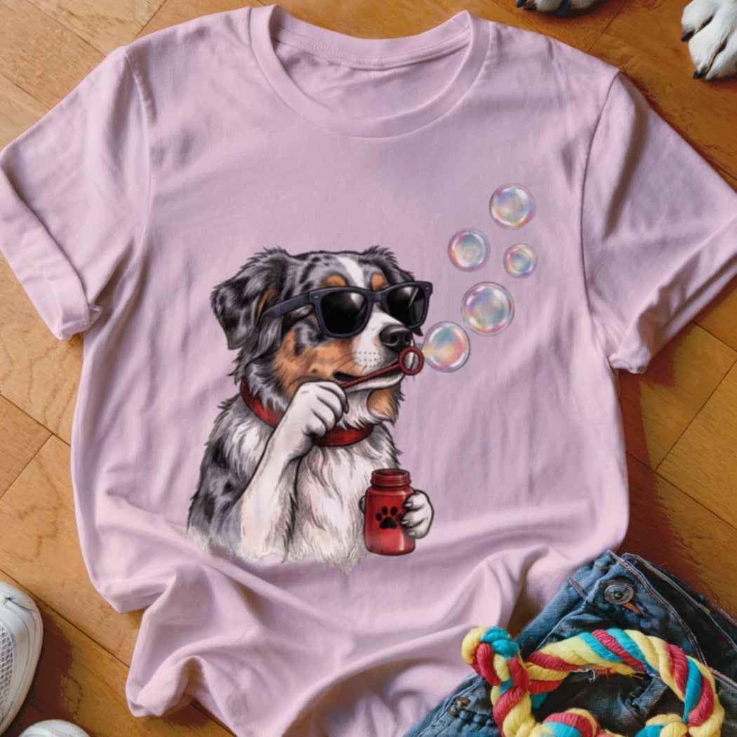 A light pink t-shirt features a graphic of an Australian Shepherd wearing sunglasses and blowing soap bubbles.